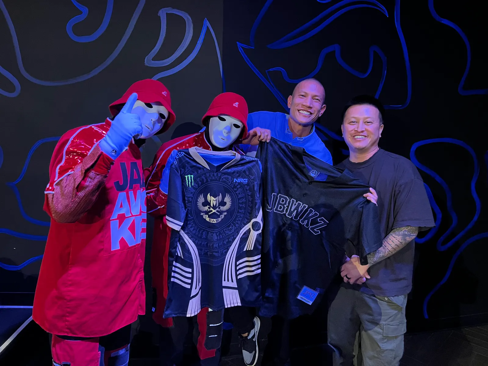 Two masked Jabbawockeez members holding a GAM Esports jersey and a JBWKZ jersey with TK Nguyen backstage in Las Vegas